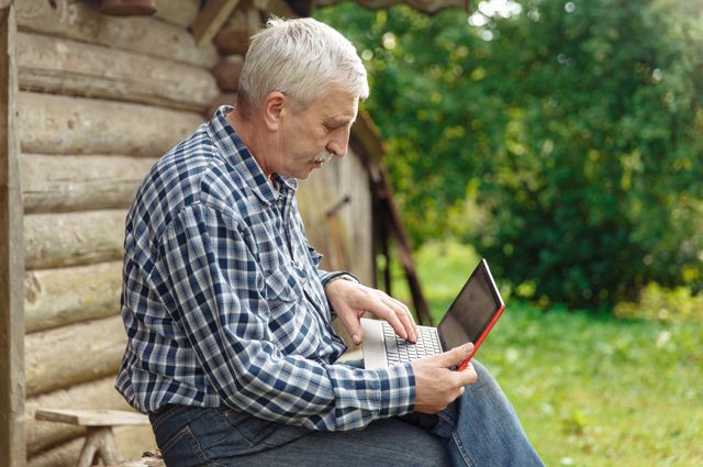 Пожилой житель села сидит за ноутбуком.