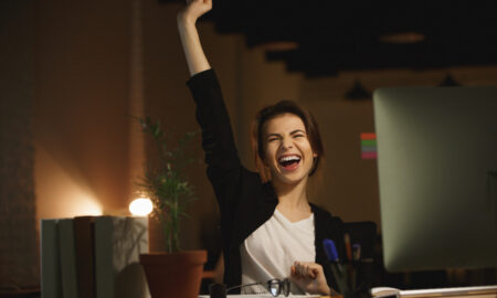 Женщина в белой футболке и чёрном пиджаке сидит за столом, улыбается и поднимает руку вверх.