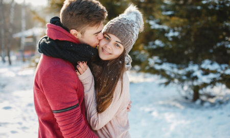 Пара обнимает друг друга на фоне заснеженного леса. Парень в красной толстовке целует девушку в щёку.