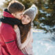 Пара обнимает друг друга на фоне заснеженного леса. Парень в красной толстовке целует девушку в щёку.