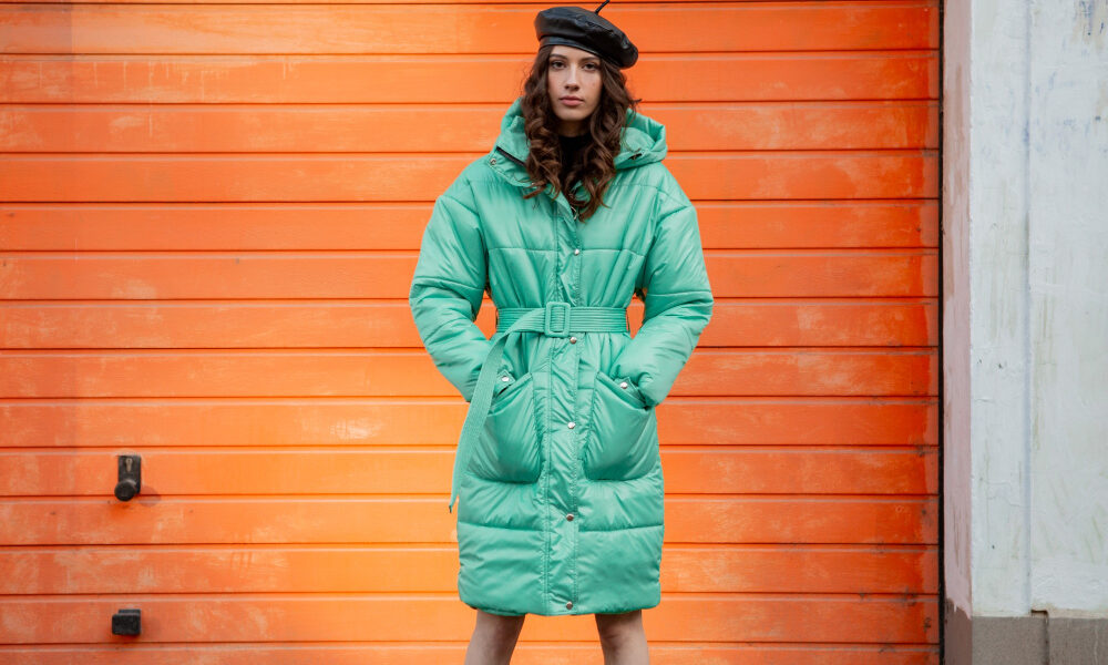 Женщина в бирюзовом пуховике на фоне оранжевой стены