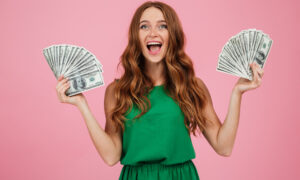 Женщина в зелёном платье на розовом фоне. Она держит веер долларов и улыбается.