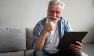 седой мужчина с планшетом в руке улыбается и показывает большой палец другой рукой