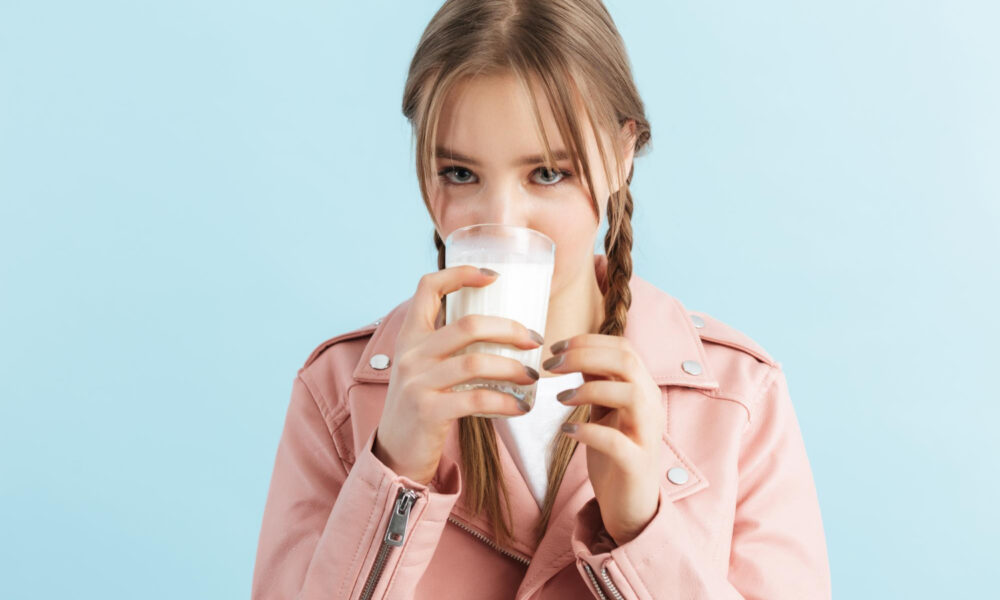 Девушка с двумя косичками, в розовой кожаной куртке и белой футболке, пьёт молоко на синем фоне