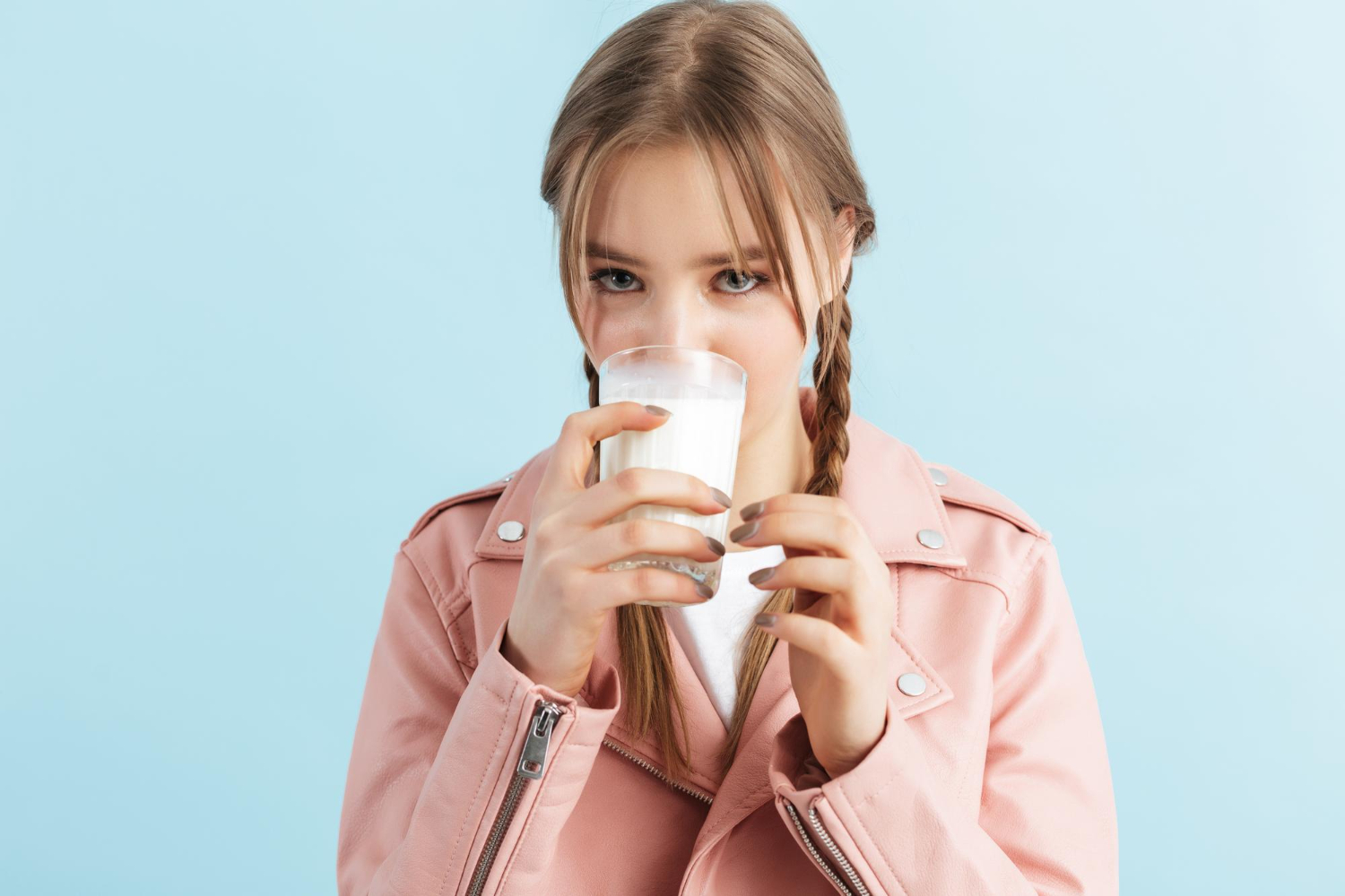 Девушка с двумя косичками, в розовой кожаной куртке и белой футболке, пьёт молоко на синем фоне