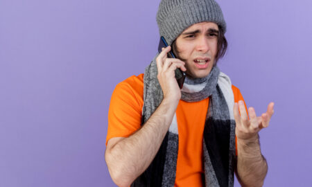 Молодой больной мужчина в зимней шапке и шарфе, смотрящий в сторону, говорит по телефону, на фиолетовом фоне.