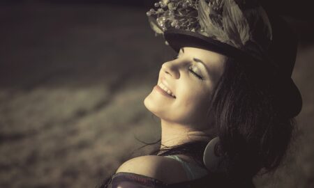 На изображении улыбающаяся женщина в шляпе с перьями. Фон размытый, тёплый свет падает на лицо женщины.