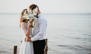 Жених и невеста целуются у воды, прикрываясь букетом.