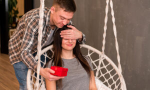 Мужчина закрыл рукой глаза девушке, которая сидит на качелях и протягивает ей подарок.