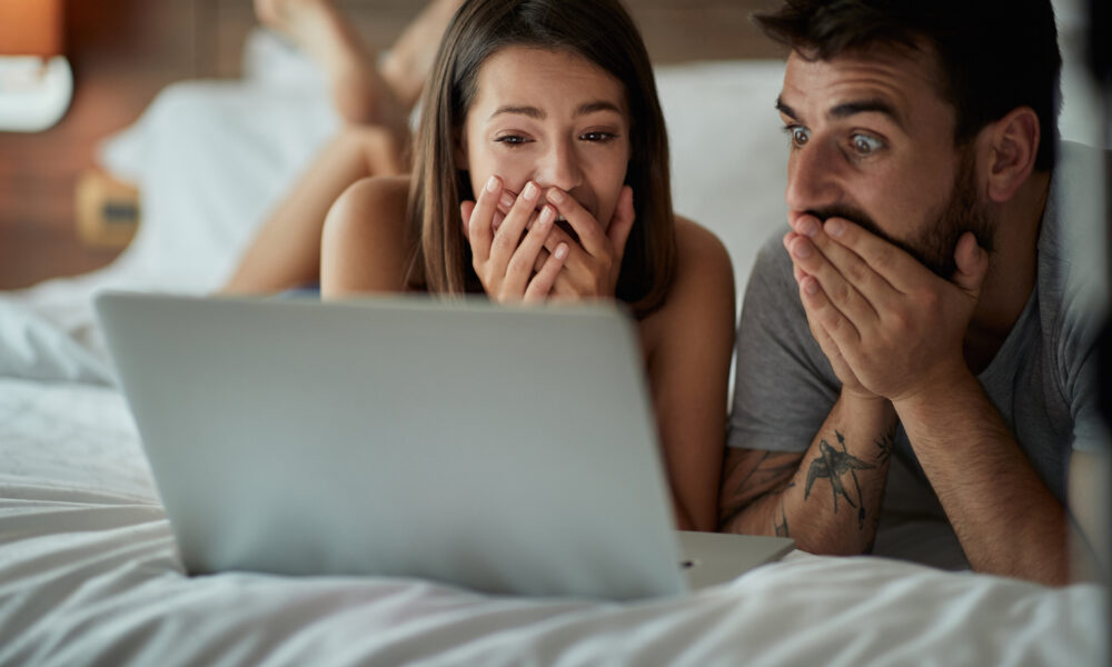 Мужчина и женщина лежат на кровати, смотрят в ноутбук и удивляются.