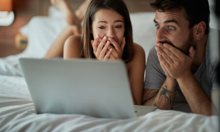Мужчина и женщина лежат на кровати, смотрят в ноутбук и удивляются.